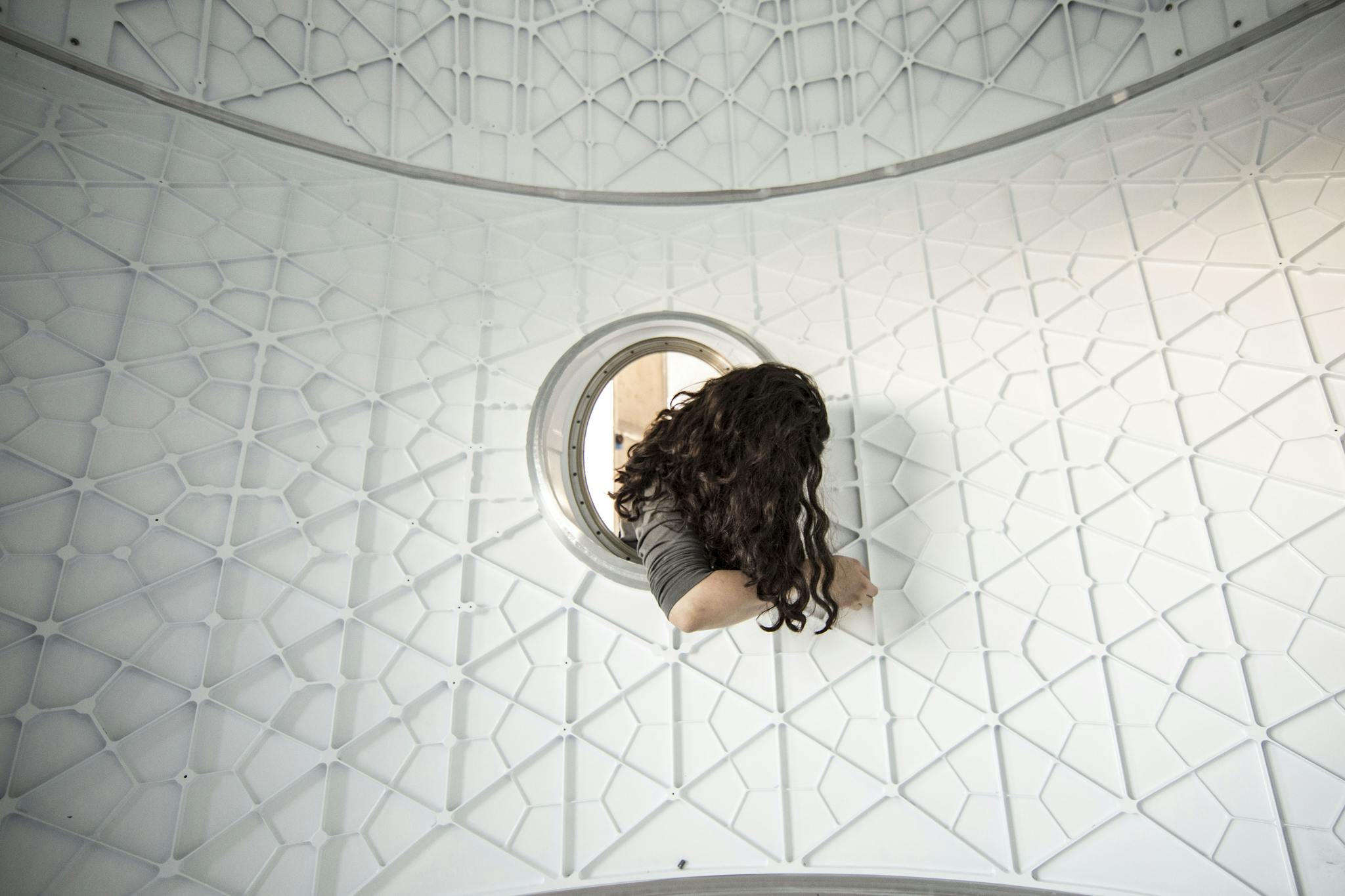 A woman peers through a circular opening in a modern geometric structure, adding a sense of curiosity.