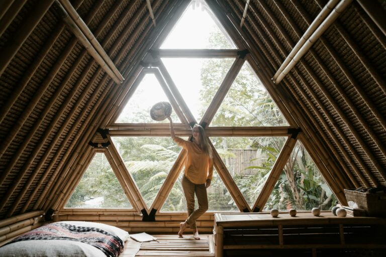Woman standing in a wooden bamboo house with triangular windows, surrounded by tropical plants.